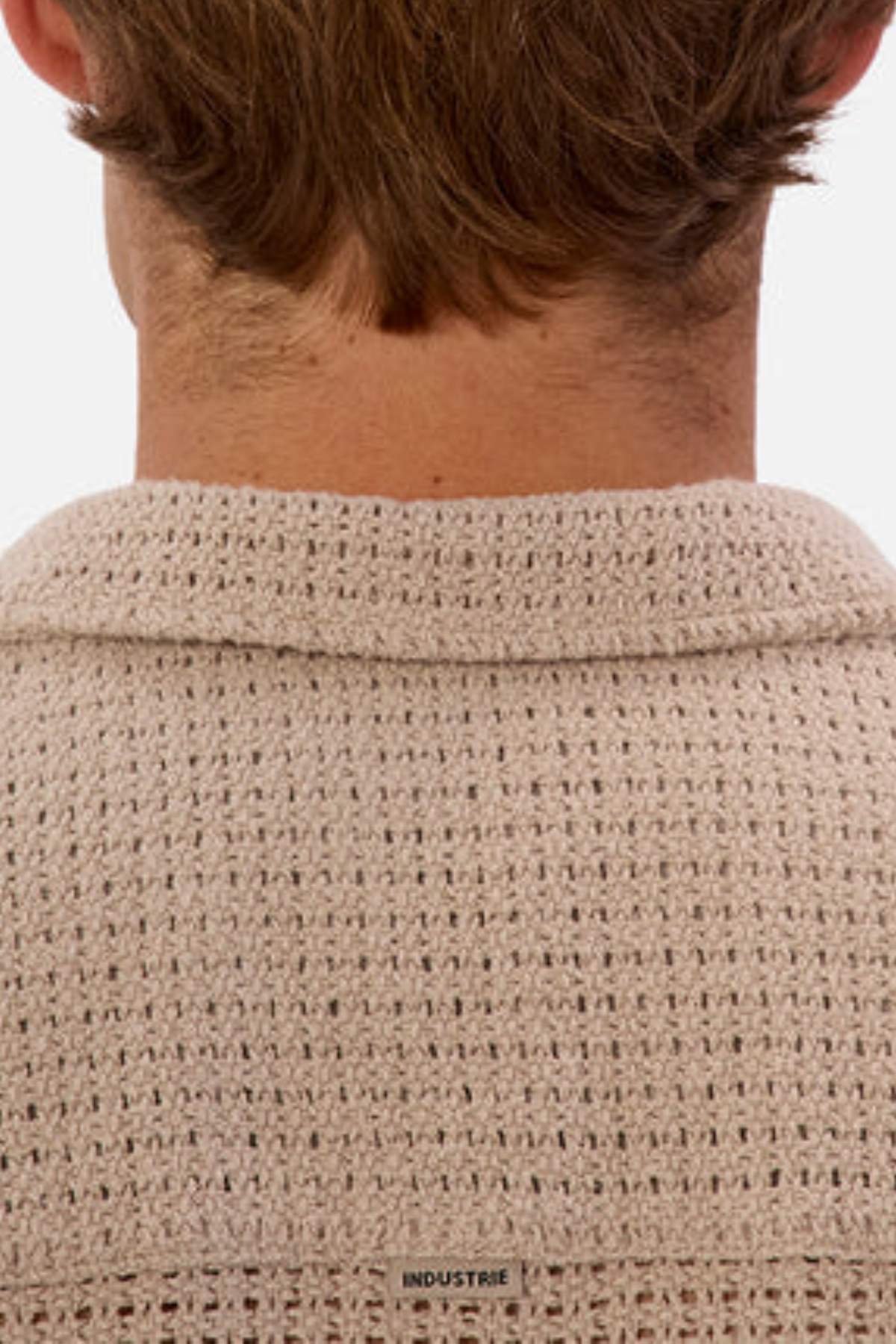 The image showcases the back of a person wearing a light brown, textured knit sweater made from breathable fabric, featuring a small "INDUSTRIE" label at the neck. The piece complements their short light brown hair and is perfect for your summer wardrobe. Product: Industrie The Egnazia Short Sleeve Shirt.
