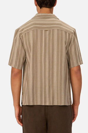 A person is shown from behind wearing the Industrie The Fabriano Short Sleeve Shirt, a beige camp collar shirt with vertical stripes and a relaxed fit, along with brown pants. The background is plain white.