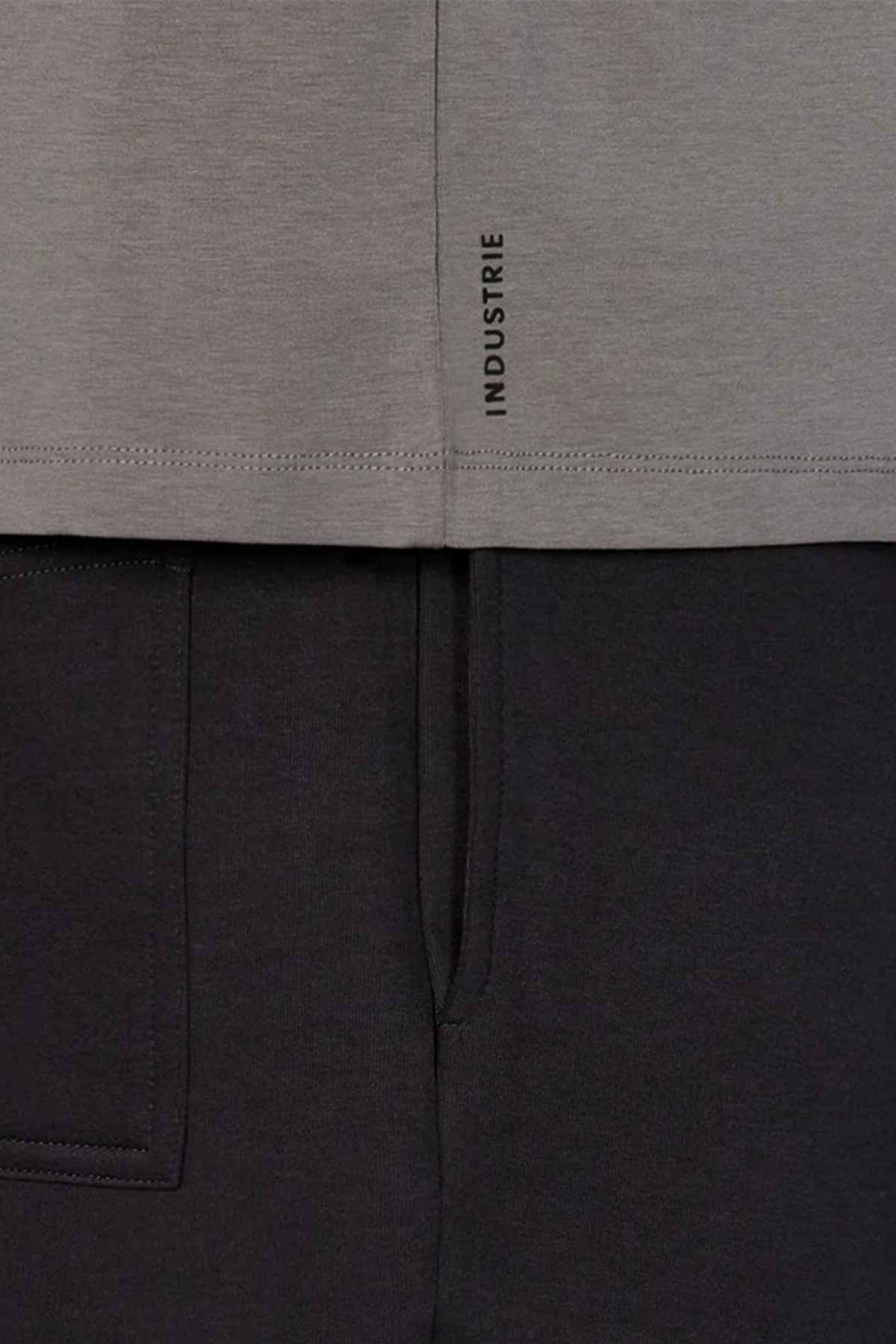 Close-up of a gray relaxed-fit Industrie The Freeman Athletic Tee featuring a small vertical "INDUSTRIE" label near the hem, styled with black pants. The image highlights the fabric textures and seams, showcasing the distinctive Industrie branding.