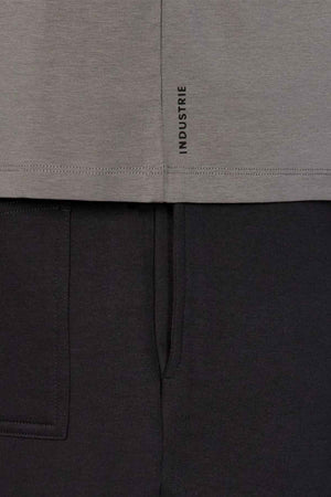 Close-up of a gray relaxed-fit Industrie The Freeman Athletic Tee featuring a small vertical "INDUSTRIE" label near the hem, styled with black pants. The image highlights the fabric textures and seams, showcasing the distinctive Industrie branding.