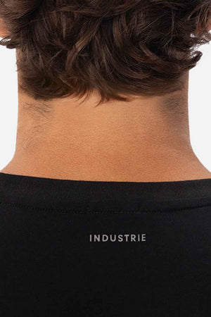 A close-up of the back of a person's neck and head highlights their dark brown hair and the upper portion of a black, relaxed-fit shirt. The word "INDUSTRIE" is elegantly printed in small white letters below the neckline, capturing the effortless style characteristic of Industrie The Freeman Athletic Tee.