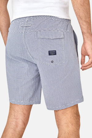 A man is shown from the waist down wearing Industrie The Freeport Bahama Short in blue and white seersucker with an elastic drawcord waist, a back pocket featuring a small navy label, and a white shirt; one hand relaxed by his side.