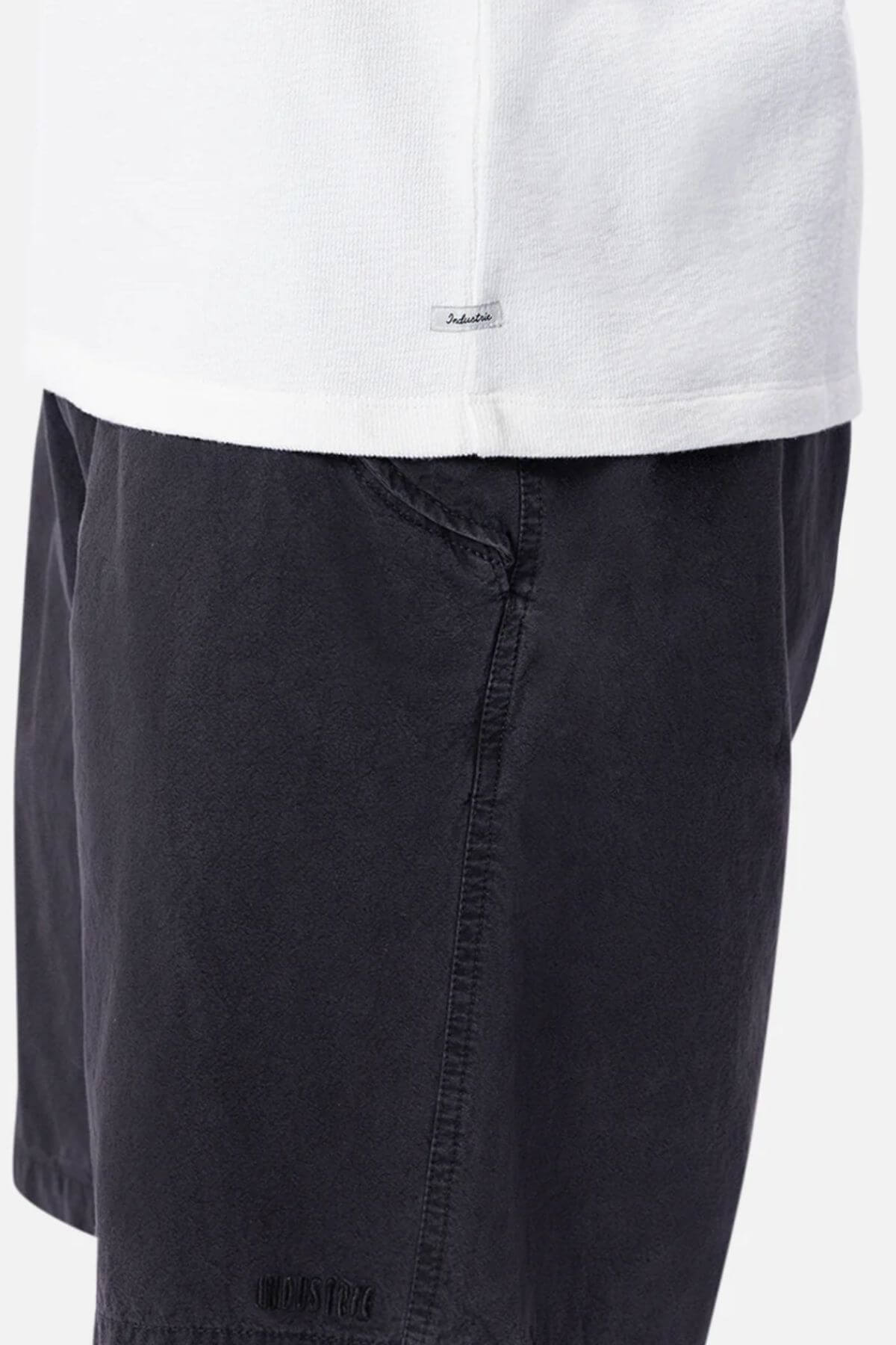 A close-up shows someone in an Industrie The Henderson Boucle Tee tucked into dark shorts. The oversized, 100% cotton tee contrasts with the shorts' subtle texture and stitched hem label.