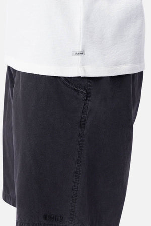 A close-up shows someone in an Industrie The Henderson Boucle Tee tucked into dark shorts. The oversized, 100% cotton tee contrasts with the shorts' subtle texture and stitched hem label.