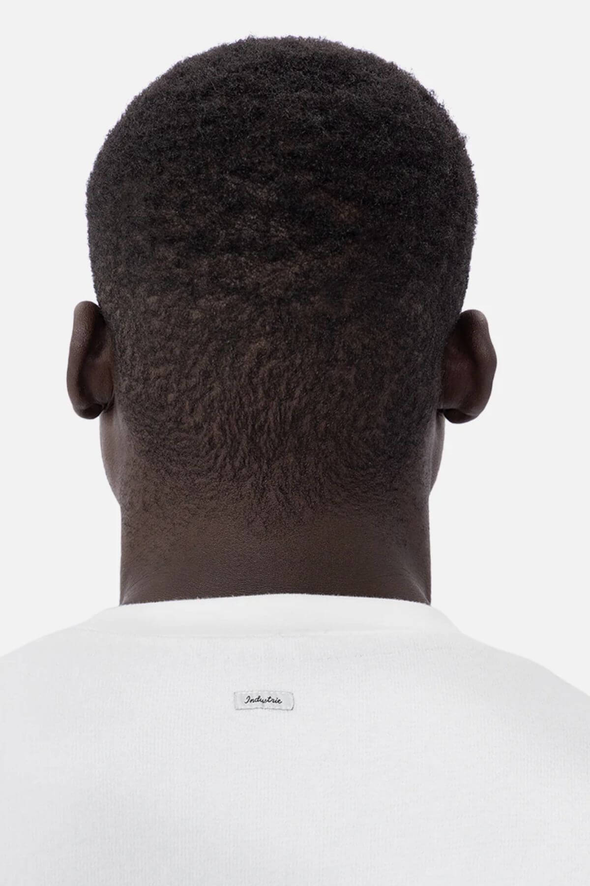 A person with short, textured hair in an oversized white shirt faces away. The plain background accentuates the small label on the back of the Industrie The Henderson Boucle Tee, made from 100% cotton.
