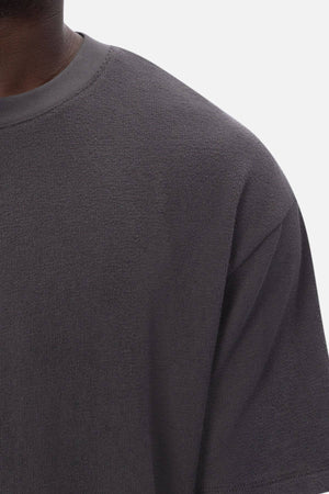 Close-up of a person wearing the Industrie The Henderson Boucle Tee, a dark gray, textured short-sleeve shirt. The shoulder, chest, and neck are visible against a plain white background.
