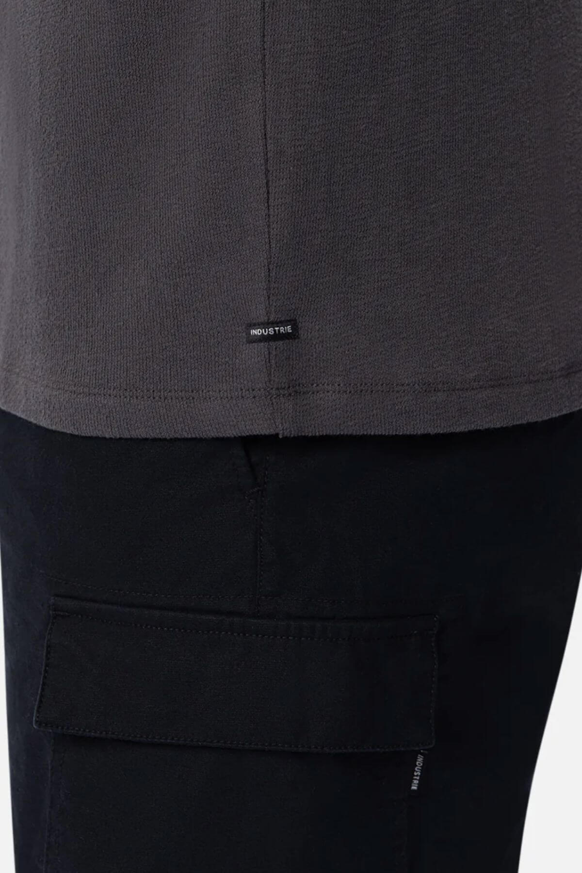 Close-up of a person wearing the Industrie The Henderson Boucle Tee in dark gray, oversized fit, paired with black flap-pocket pants. A small black "INDUSTRIE" tag is sewn near the shirt’s bottom hem.