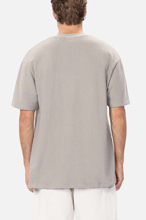 An individual with short hair is wearing an oversized, light gray Industrie The Henderson Boucle Tee (100% cotton) paired with white pants. Facing away from the camera, they showcase the relaxed back view of the outfit against a plain white background.