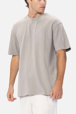 A man wearing the Industrie The Henderson Boucle Tee—a loose-fitting, short-sleeve, light gray shirt—paired with white pants stands against a plain background, looking to his left.