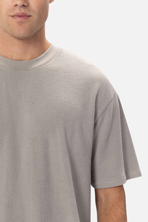 A partial view shows someone in an Industrie The Henderson Boucle Tee, featuring its oversized fit in plain gray 100% cotton. Their left face is partially hidden against a plain, light background.
