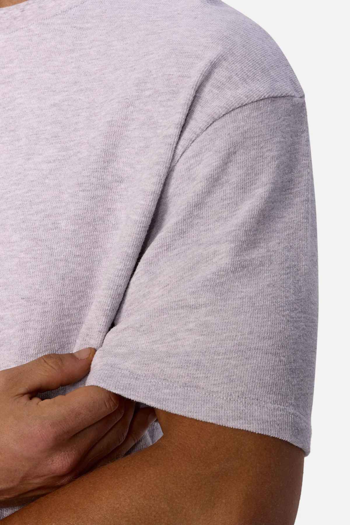 A person wears the Industrie The Henderson Boucle Tee in an oversized style, featuring short sleeves and a light gray color, as they gently hold their elbow with their right hand. The image highlights the upper section of this 100% cotton t-shirt, focusing on its texture against a plain white background.