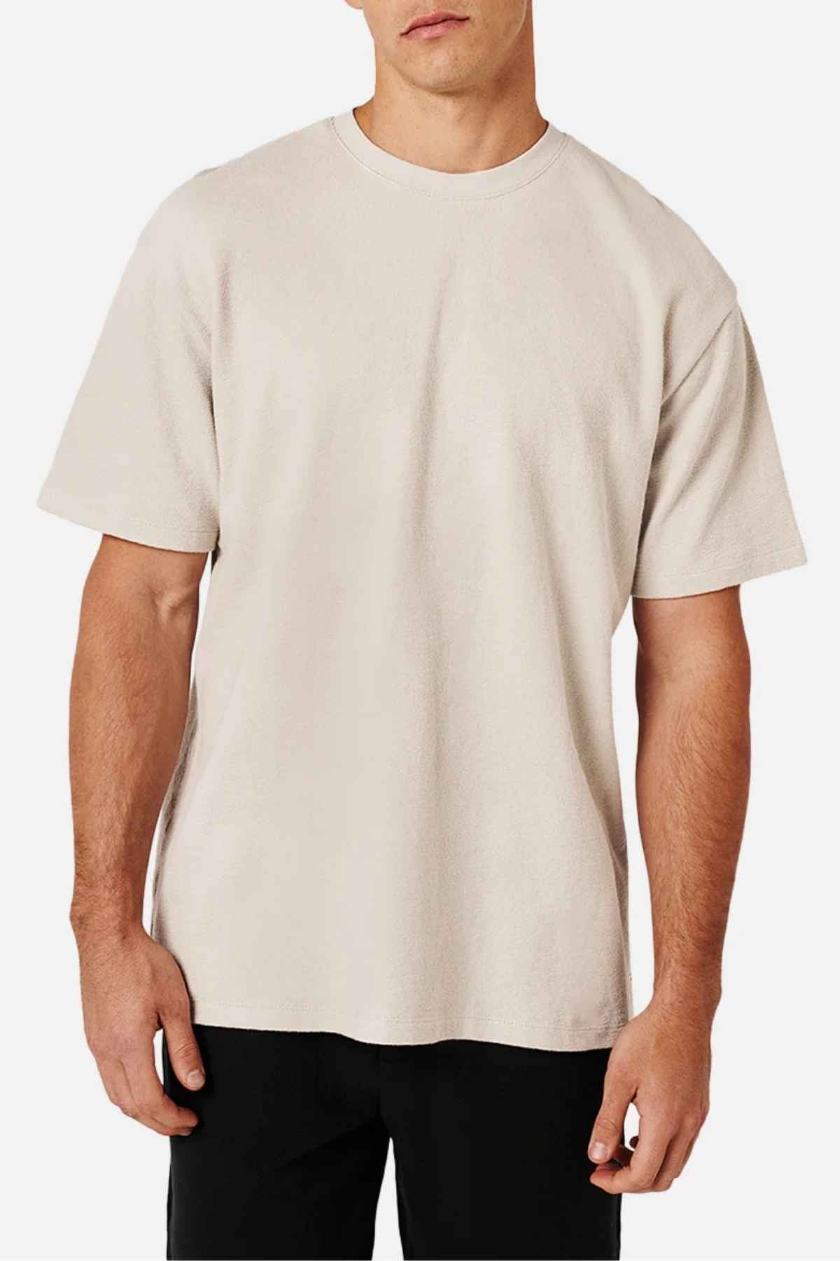 A person is wearing the Industrie The Henderson Boucle Tee, an oversized beige T-shirt made of 100% cotton, featuring short sleeves and a crew neckline. They have paired it with black pants and are standing against a white background.