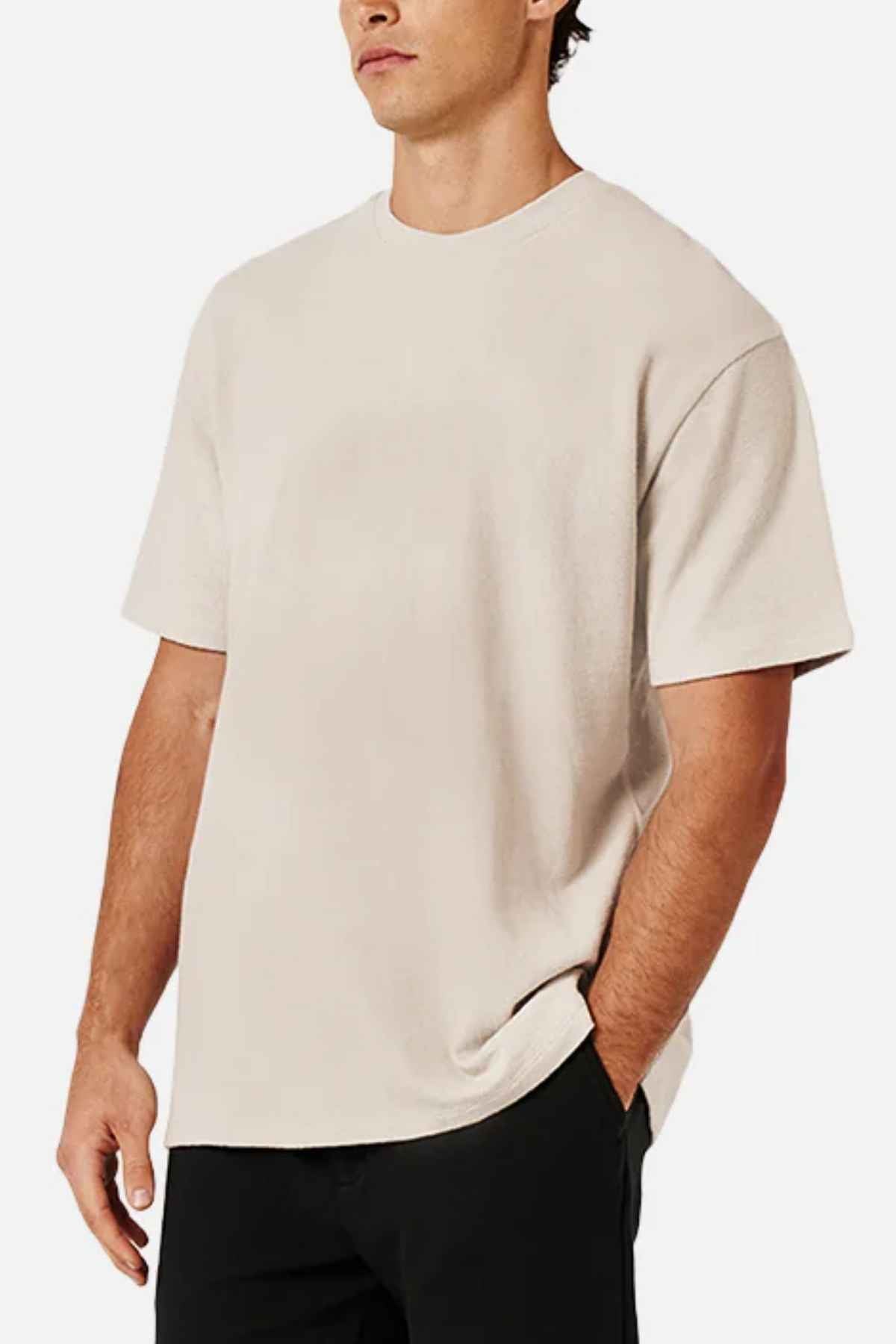 A man in an oversized beige Industrie The Henderson Boucle Tee, made from 100% cotton, and black pants stands against a plain white background.