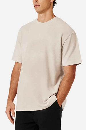 A man in an oversized beige Industrie The Henderson Boucle Tee, made from 100% cotton, and black pants stands against a plain white background.