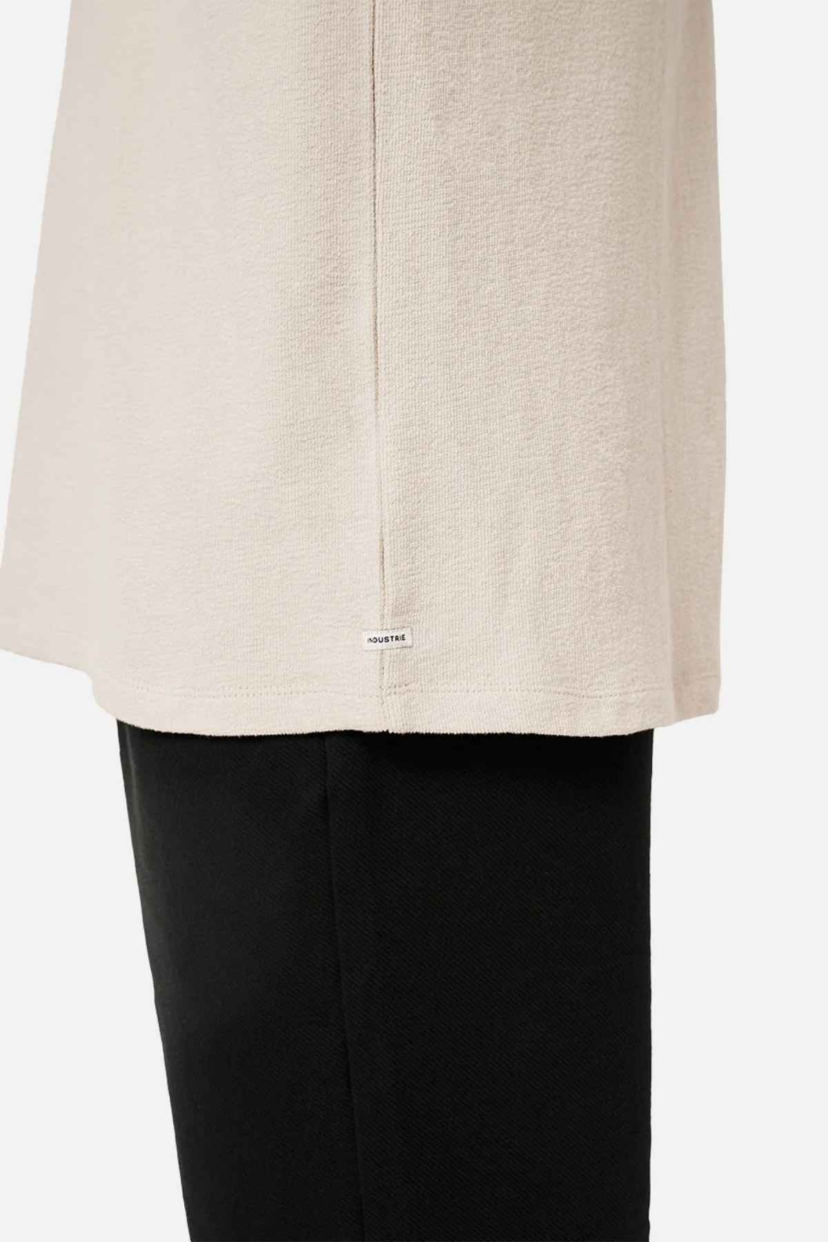 Close-up of a person wearing the Industrie The Henderson Boucle Tee in beige paired with black pants. The focus is on the side seam, highlighting a small white tag with text. Made from 100% cotton, this tee provides an effortlessly oversized look against the plain white backdrop.