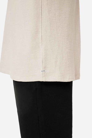 Close-up of a person wearing the Industrie The Henderson Boucle Tee in beige paired with black pants. The focus is on the side seam, highlighting a small white tag with text. Made from 100% cotton, this tee provides an effortlessly oversized look against the plain white backdrop.
