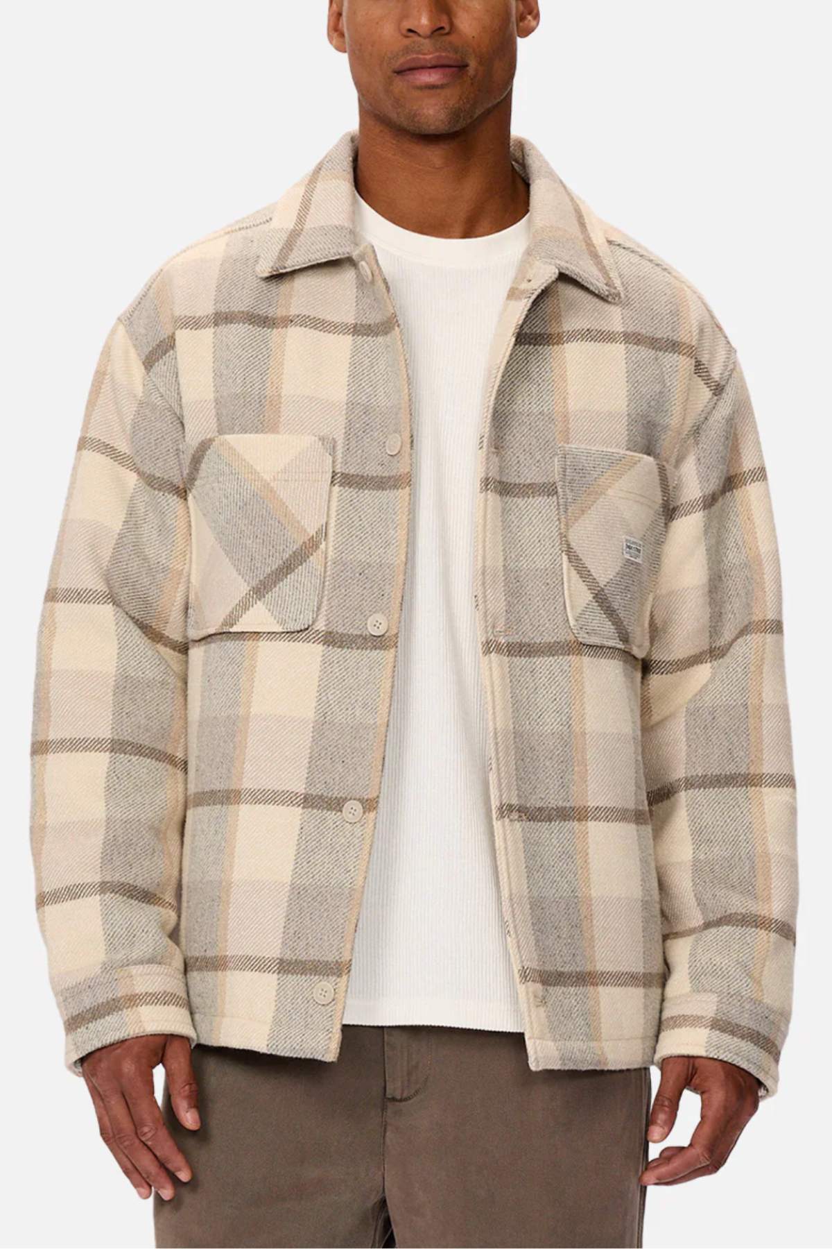Sporting the Industrie The Hillside Jacket, a beige and gray check pattern button-up featuring two chest pockets over a crisp white shirt, teamed with brown pants on a plain white backdrop, this ensemble perfectly melds style and comfort.