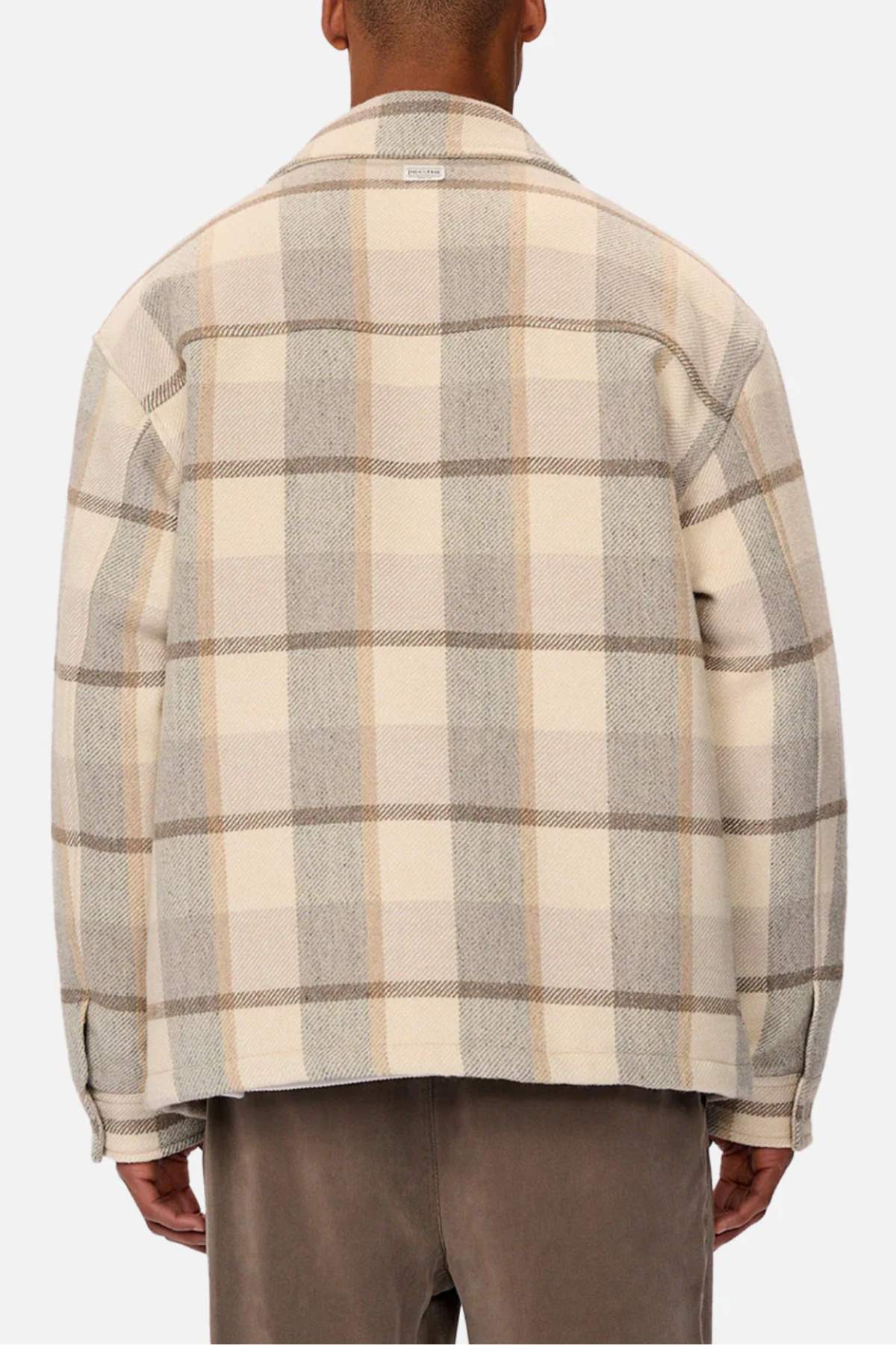 A person is seen from behind wearing the Industrie The Hillside Jacket, displaying its large plaid check in beige, cream, and gray. It is stylishly paired with brown pants against a white background.
