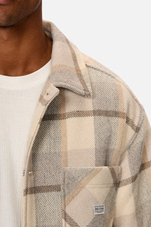 A person is wearing the Industrie The Hillside Jacket, displaying a plaid beige and grey check pattern over a white ribbed shirt. The jacket's top button is undone, revealing a "Industrie" label on the pocket. Focus is on the upper torso against a plain background.