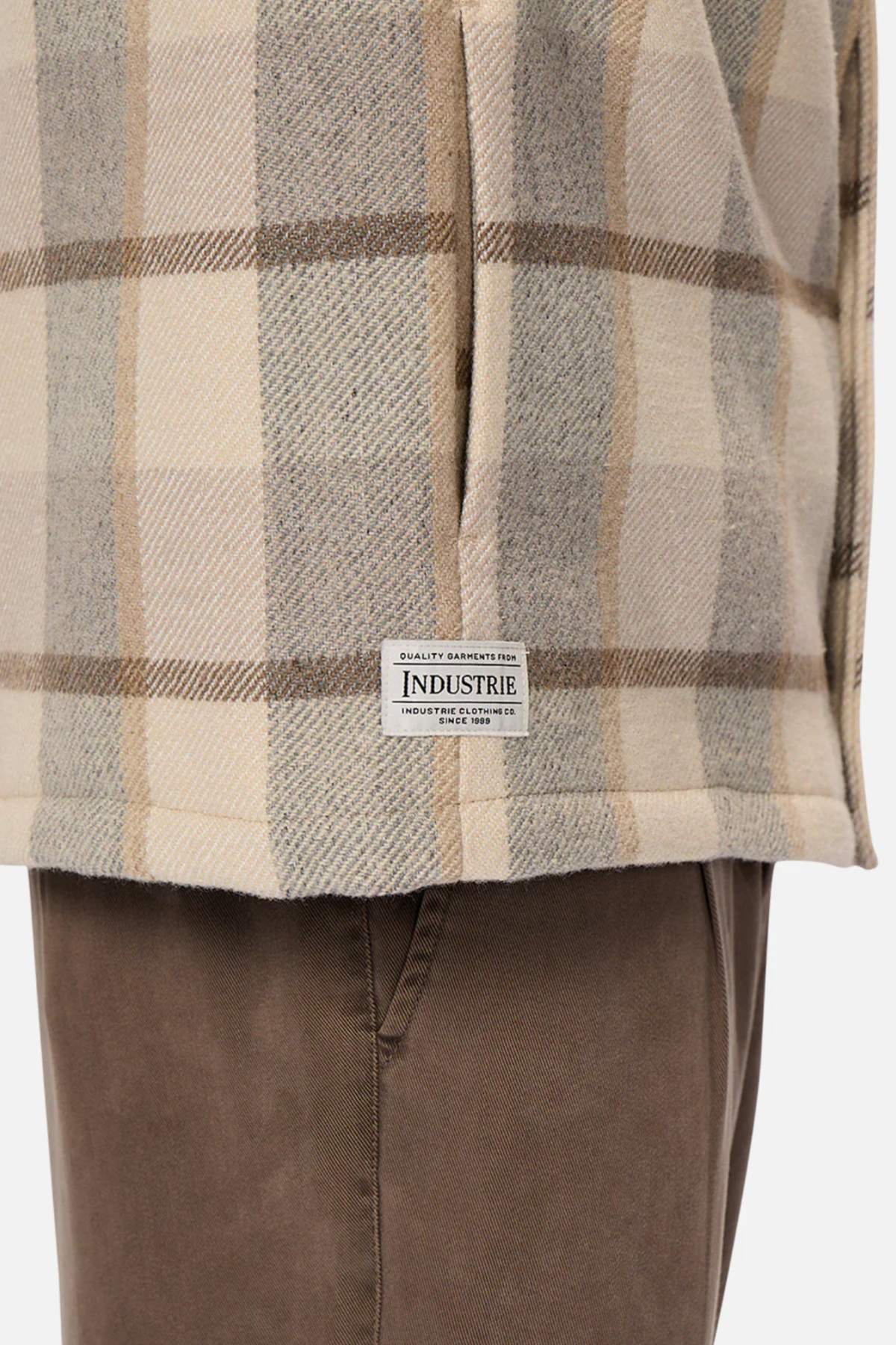 A person wears a soft, insulating beige and gray checkered shirt over brown pants, with an "Industrie" label. The flowing hem resembles the Industrie The Hillside Jacket.