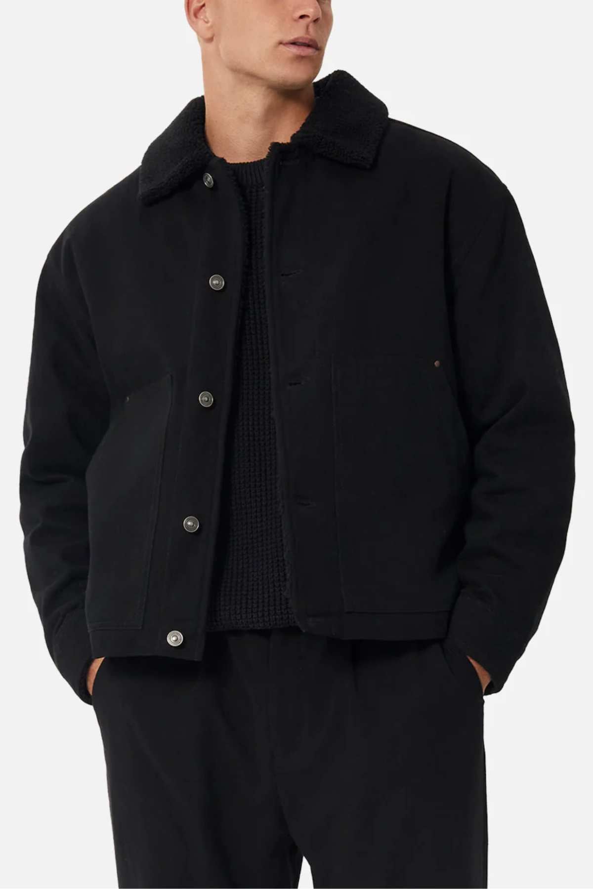 Someone stands with hands in their pockets, wearing the Industrie The Huntsville Jacket with a classic collar and sherpa lining, layered over a black sweater and black pants.