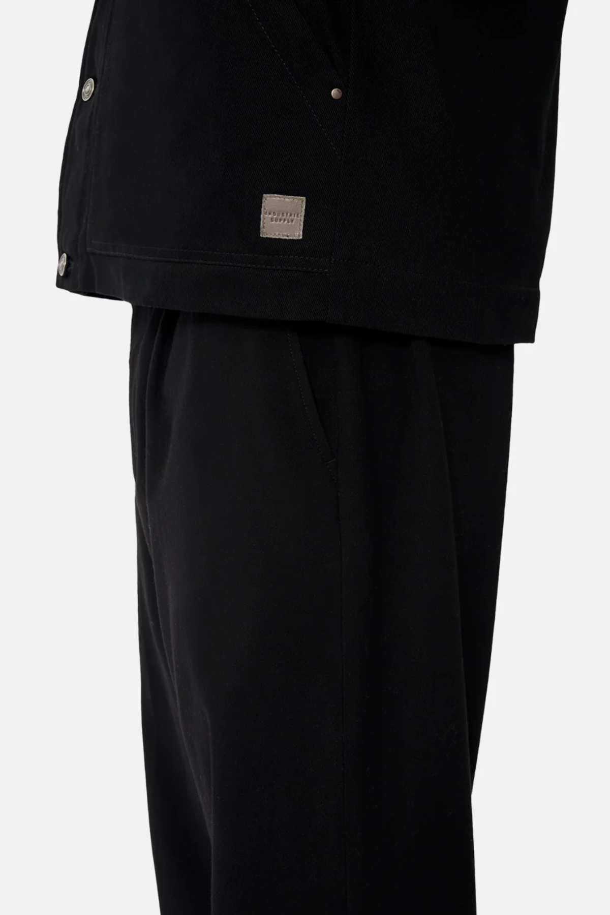 A close-up of a person in black pants and the Industrie The Huntsville Jacket, which has metal buttons, a classic collar, sherpa lining, and a small rectangular fabric label by the hem. Their left hand is in their pocket.