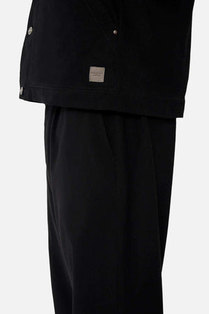 A close-up of a person in black pants and the Industrie The Huntsville Jacket, which has metal buttons, a classic collar, sherpa lining, and a small rectangular fabric label by the hem. Their left hand is in their pocket.