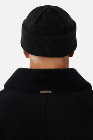 The image displays the back of a person in the Industrie The Huntsville Jacket with a classic collar and textured finish, plus a small metallic tag at the center. They're also wearing a black knit beanie against a light background.