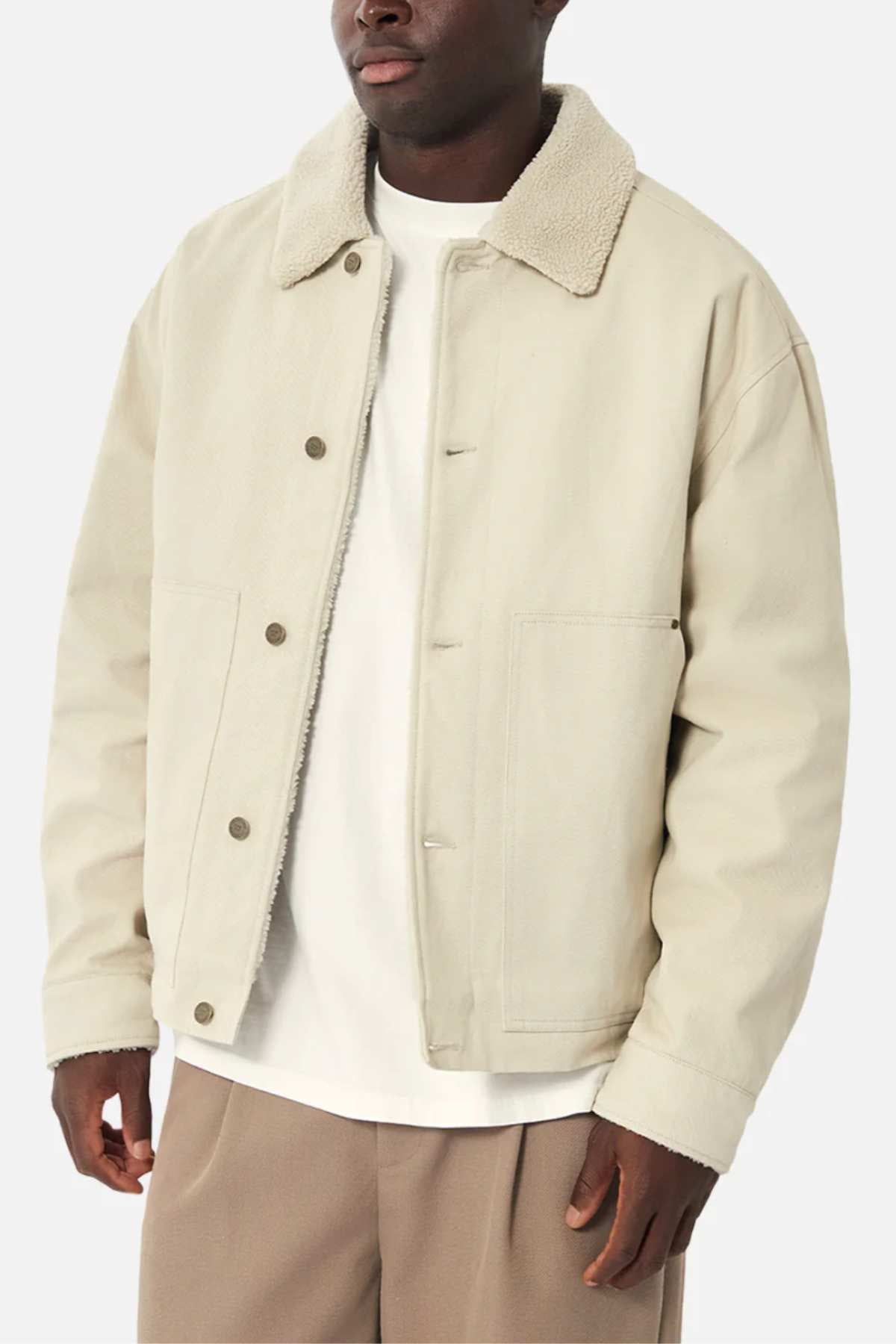 A man wears the Industrie The Huntsville Jacket in beige with a sherpa-lined classic collar over a white shirt and brown pants, creating a stylish look against a simple backdrop.