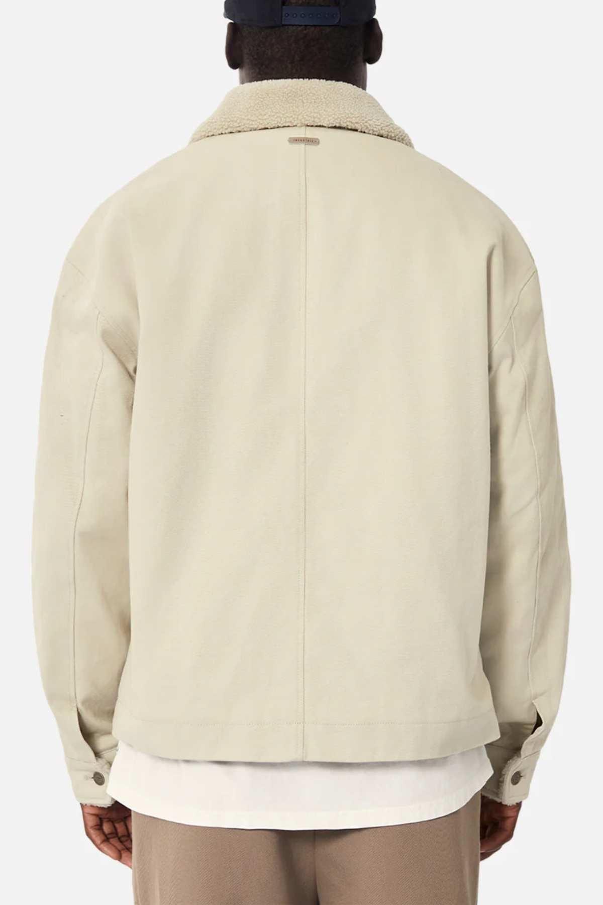 Seen from behind, a person wears Industrie The Huntsville Jacket in beige with a white collar over a white shirt, paired with a navy cap. The crisp backdrop accentuates the jacket's timeless appeal.