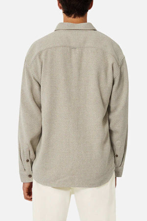 The individual is seen from the back wearing a light gray, relaxed-fit long-sleeved shirt with a subtle herringbone pattern. This is the Industrie The Jackson Shirt, complete with a collar and buttoned cuffs, paired stylishly with white pants against a plain white background.