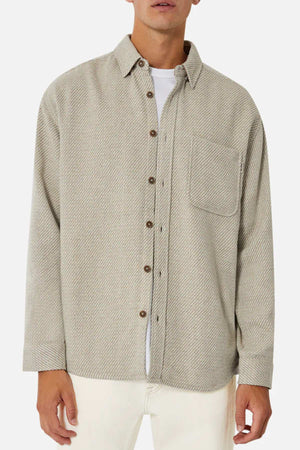 A man wears the Industrie The Jackson Shirt in light gray with a subtle herringbone pattern, featuring a chest pocket and brown buttons, over a white T-shirt. This stylish long-sleeved shirt is paired with light-colored pants for a relaxed fit.