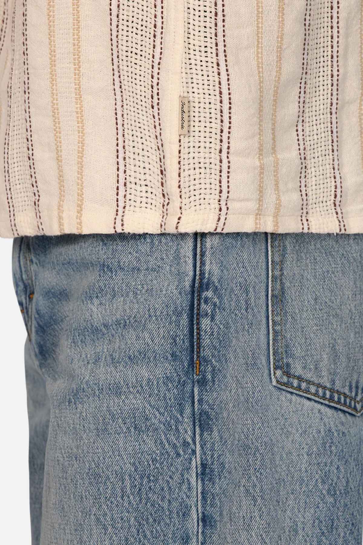 Close-up of a person wearing light blue denim jeans and the Industrie The Lakeville Short Sleeve Shirt in beige, featuring vertical stripes and a textured stripe design. The focus is on the seam and pocket details of the jeans and the hem of this stylish linen viscose blend shirt.
