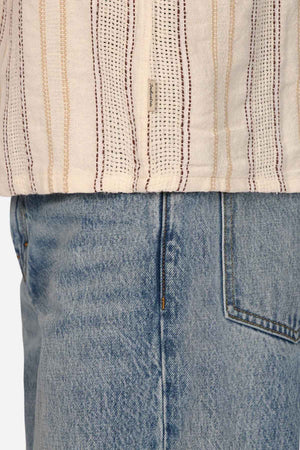 Close-up of a person wearing light blue denim jeans and the Industrie The Lakeville Short Sleeve Shirt in beige, featuring vertical stripes and a textured stripe design. The focus is on the seam and pocket details of the jeans and the hem of this stylish linen viscose blend shirt.