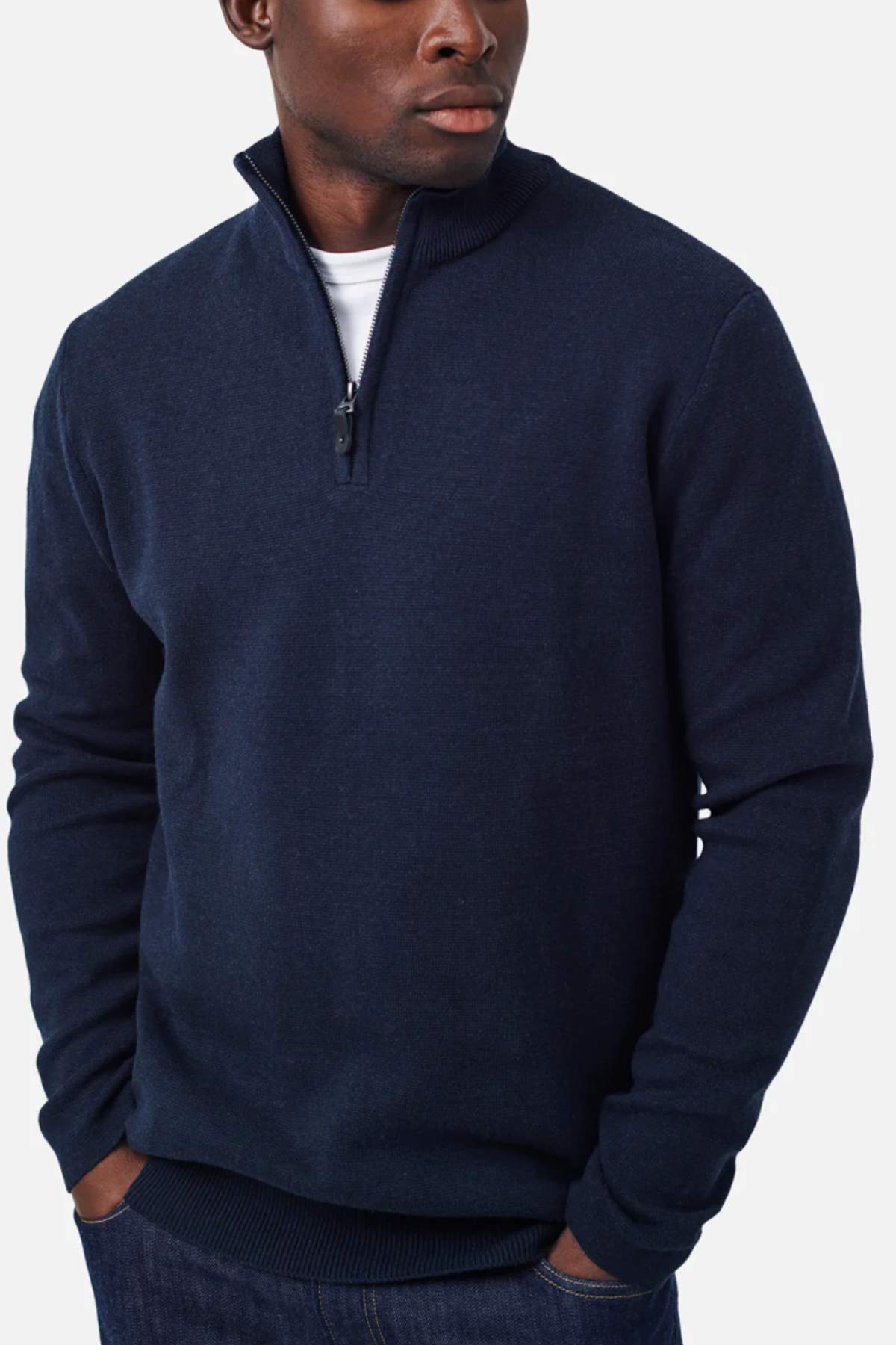 A man is wearing the Industrie The Lakewood Zip Neck, a regular fit navy blue sweater, with a white t-shirt underneath. He has his hands in the pockets of his dark blue jeans and is looking to the side. The background is plain white.
