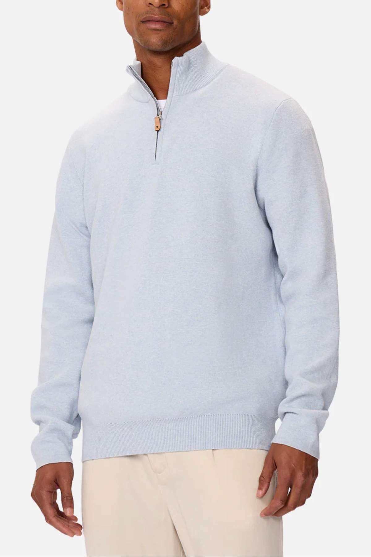 A person in the Industrie The Lakewood Zip Neck, a light blue cotton jumper with a zip neck, and beige pants stands against a white background. The high collar and brown zipper pull add detail, while the regular fit allows their hands to rest comfortably at their sides.