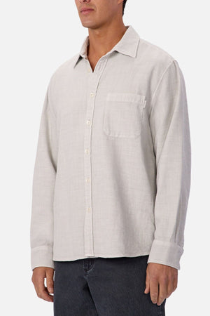 A man wears the Industrie Lisbon Long Sleeve Shirt in light beige—100% cotton, classic collar, and chest pocket—paired with dark pants, standing against a plain white background.