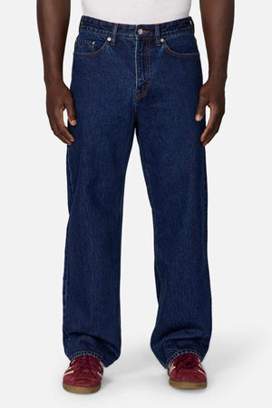 A person standing against a white background is wearing the Industrie The Lone Star Jean, a pair of dark blue, loose-fitting jeans made from 100% cotton denim. The jeans feature a classic five-pocket style with visible stitching. They complete the look with red sneakers and a white shirt, hands resting at their sides.