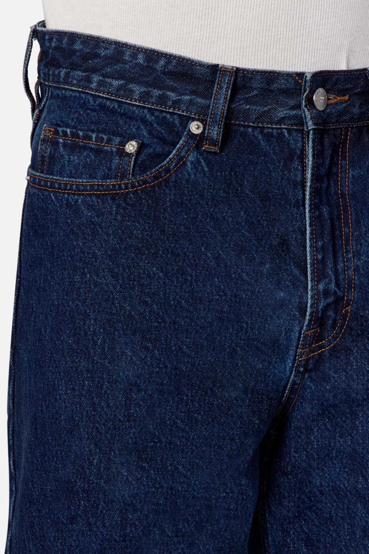 Close-up of a person's waist wearing the Industrie The Lone Star Jean in dark blue denim with orange stitching. The image highlights the front side of these baggy fit jeans, showcasing details such as the button, top of the zipper, front pocket, and belt loops. A white shirt is partially visible above these 100% cotton denim jeans.
