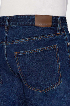Close-up of the back of a person wearing Industrie The Lone Star Jean in dark blue 100% cotton denim with visible stitching and a single back pocket on the right side. A brown leather patch with the word "INDUSTRIE" is sewn on the waistband. The person, wearing a white shirt tucked in, appears to be sporting these jeans.