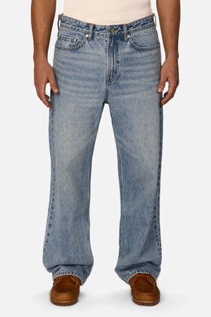 A person is wearing a pair of Industrie The Lone Star Jean in light blue with a loose fit and straight-leg cut, featuring visible seams and a slight fade. They are also wearing brown moccasin-style shoes. Their hands rest by their sides, revealing the waistband of a partially visible white shirt.
