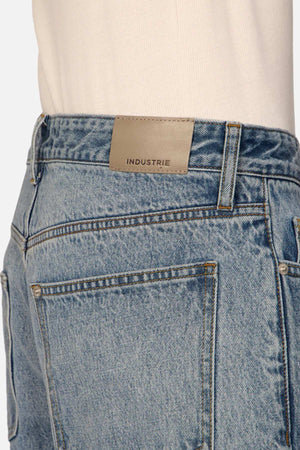Close-up of a person wearing Industrie The Lone Star Jean in light blue denim, featuring a leather patch on the back waistband labeled "INDUSTRIE." The person is also wearing a light-colored shirt tucked into the jeans. The focus is on the back pocket and the brand label.