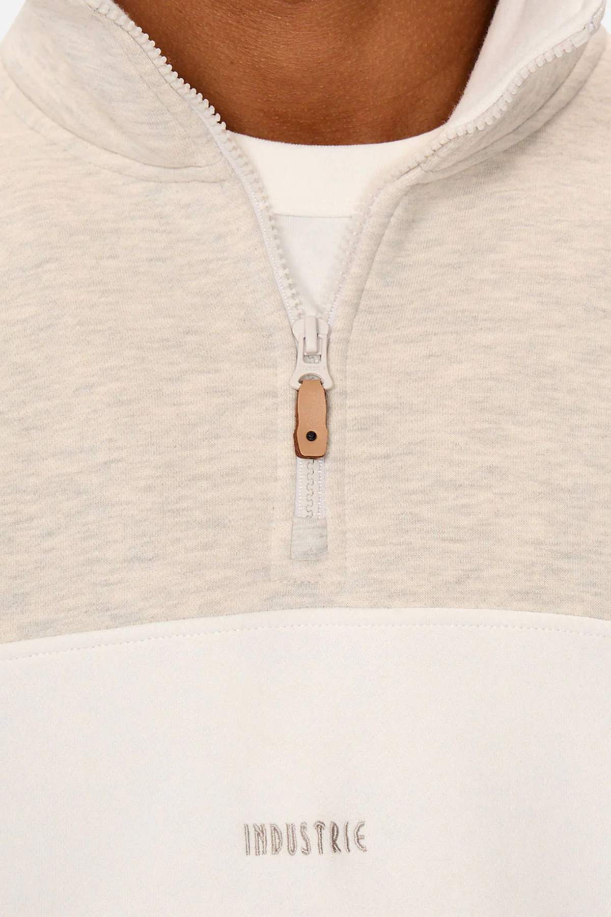 Close-up of a person wearing Industrie The Luton Track Top, showcasing a light-colored quarter-zip pullover with a white undershirt. It features "INDUSTRIE" embroidered on the chest, soft fleece lining, and a brown zipper pull.