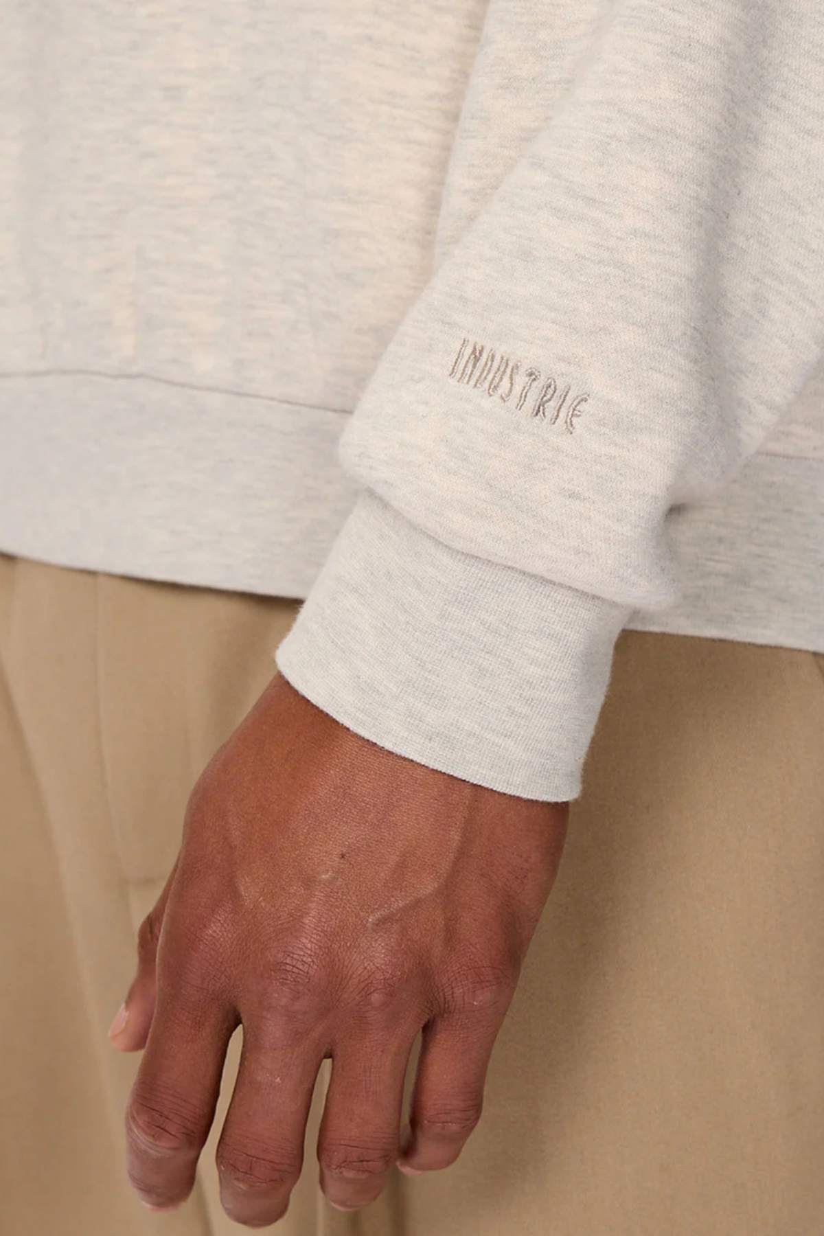 Close-up of a hand and arm wearing the Industrie The Luton Track Top in light gray, featuring "INDUSTRIE" embroidered on the sleeve cuff. The quarter-zip neck adds style, and its soft fleece lining ensures comfort. They pair it with beige pants against a neutral background.