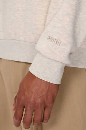 Close-up of a hand and arm wearing the Industrie The Luton Track Top in light gray, featuring "INDUSTRIE" embroidered on the sleeve cuff. The quarter-zip neck adds style, and its soft fleece lining ensures comfort. They pair it with beige pants against a neutral background.