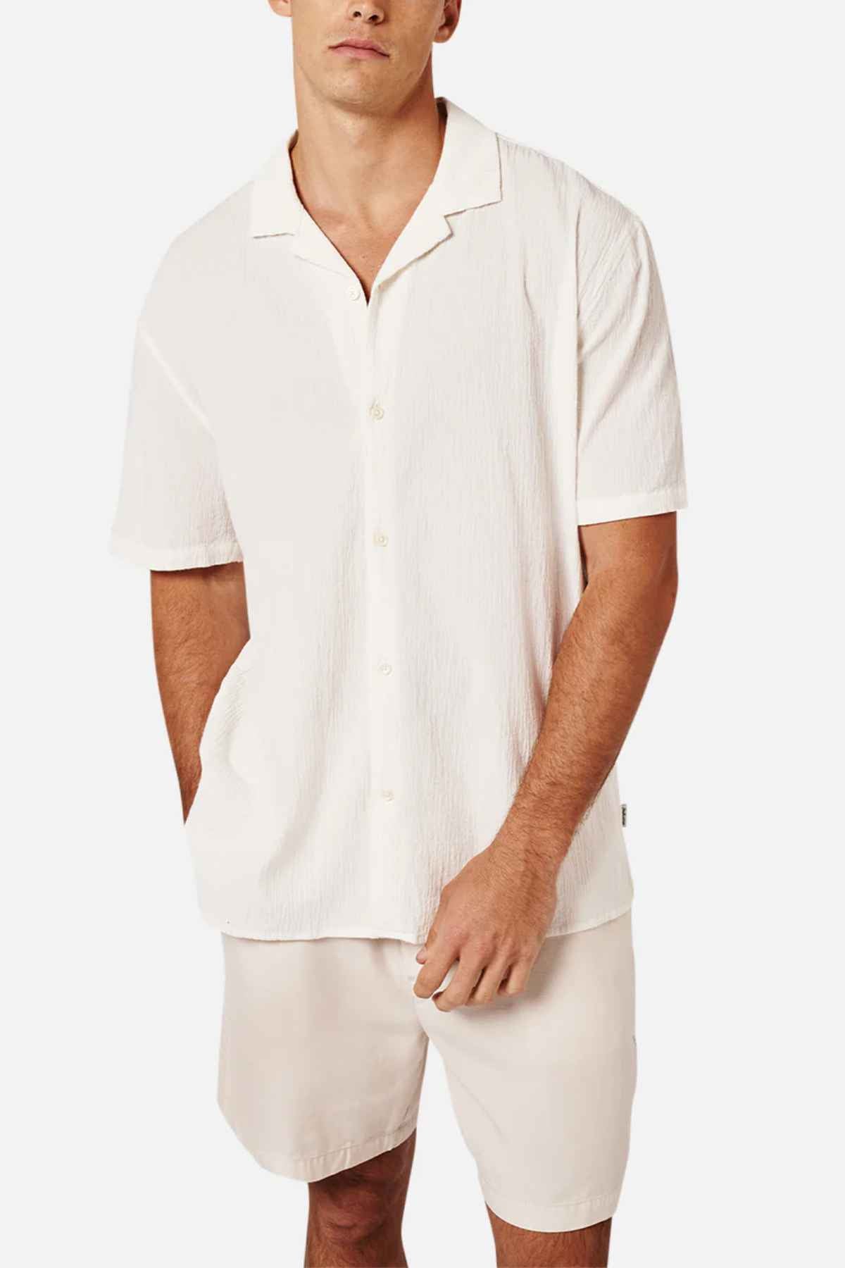 A man is wearing the Industrie The Magna Short Sleeve Shirt, which has a cream color and an oversized, short-sleeve design. He pairs it with matching cream shorts. The Summer shirt features a relaxed fit and textured, breathable cotton fabric. One of his hands rests in his pocket against a plain white background.