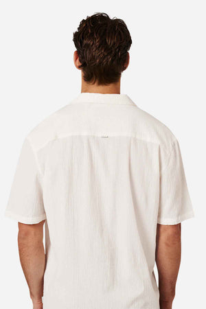 A person with short, wavy hair faces away from the camera, displaying the back view of an oversized fit, white "Industrie The Magna Short Sleeve Shirt" made of breathable cotton with a pointed collar. The background is plain white.