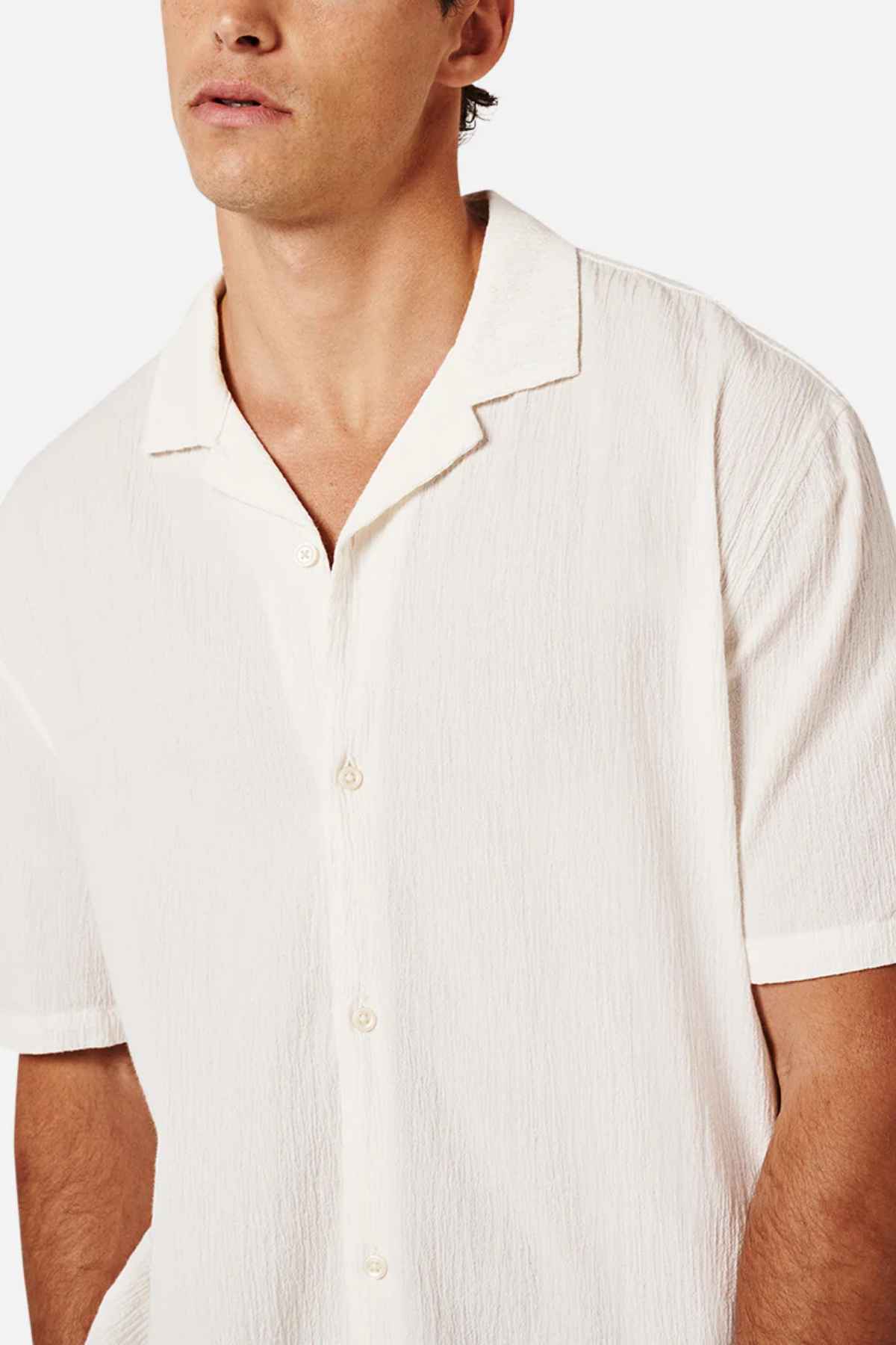 A man is shown from the shoulders up, wearing the Industrie The Magna Short Sleeve Shirt. This white textured button-up summer shirt features breathable cotton fabric and an oversized fit, perfectly complementing his short dark hair and partly out-of-frame face against a plain white background that highlights the shirt's texture and light color.