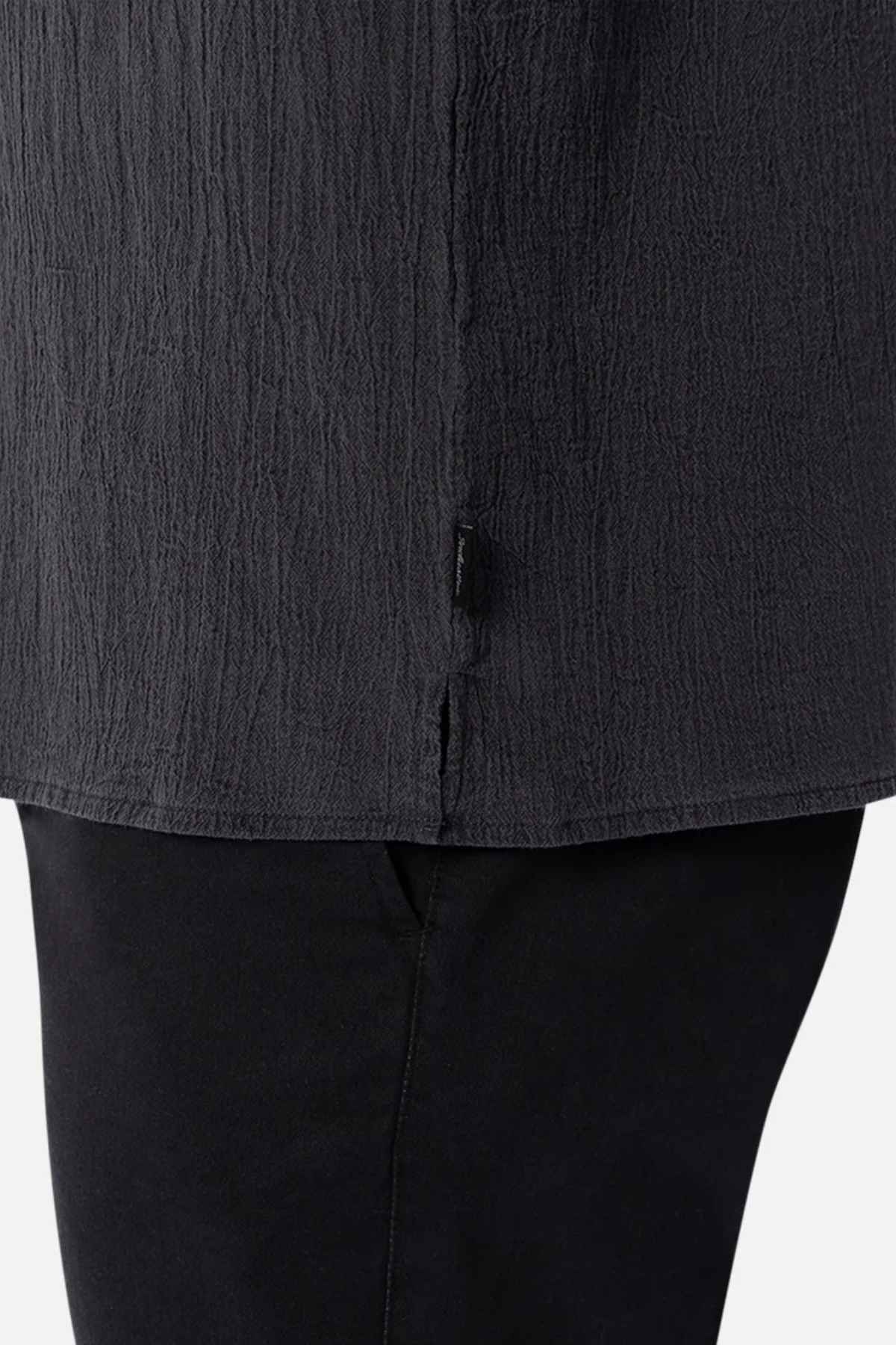 Close-up of a black crinkled fabric showcasing a hemline and a small tag. The garment, the oversized Industrie The Magna Short Sleeve Shirt, is set against a white background.