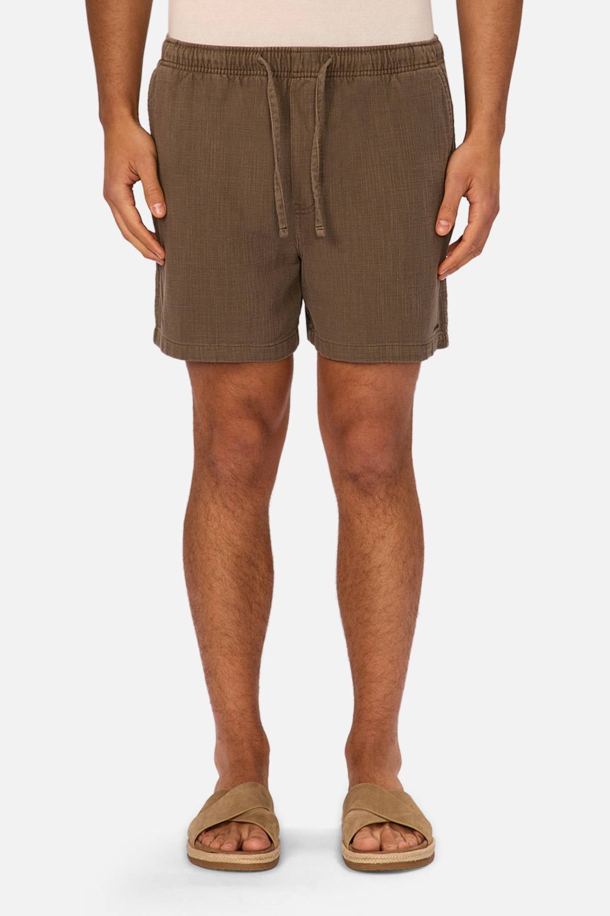 A person is seen from the waist down wearing Industrie The Mallorca Short, made of lightweight textured fabric with an elasticated waist, paired with tan slide sandals, standing against a plain white background.